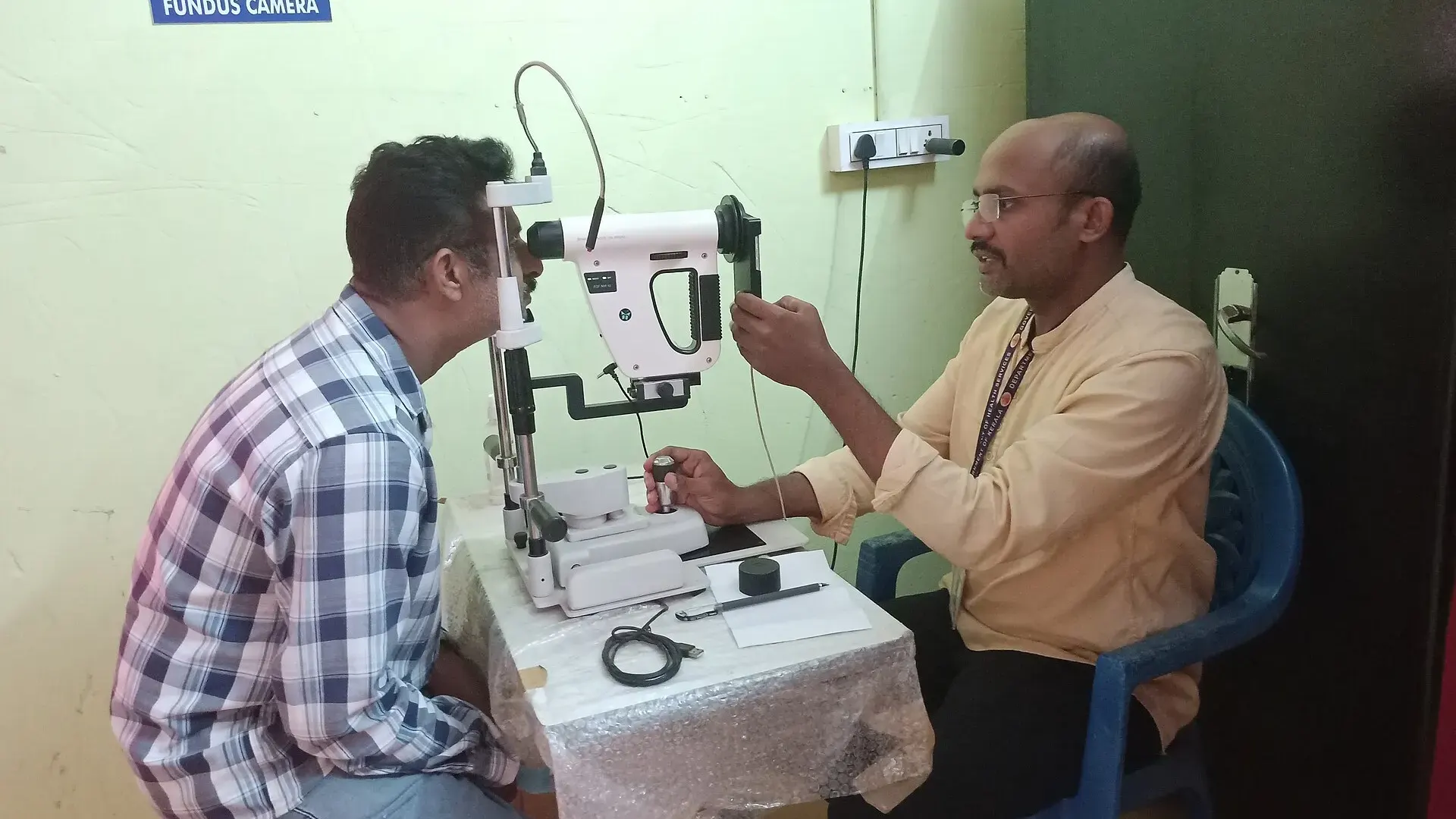 A man rests his chin on a non-mydriatic fundus camera while a health worker photographs the back of his eye in a clinic.