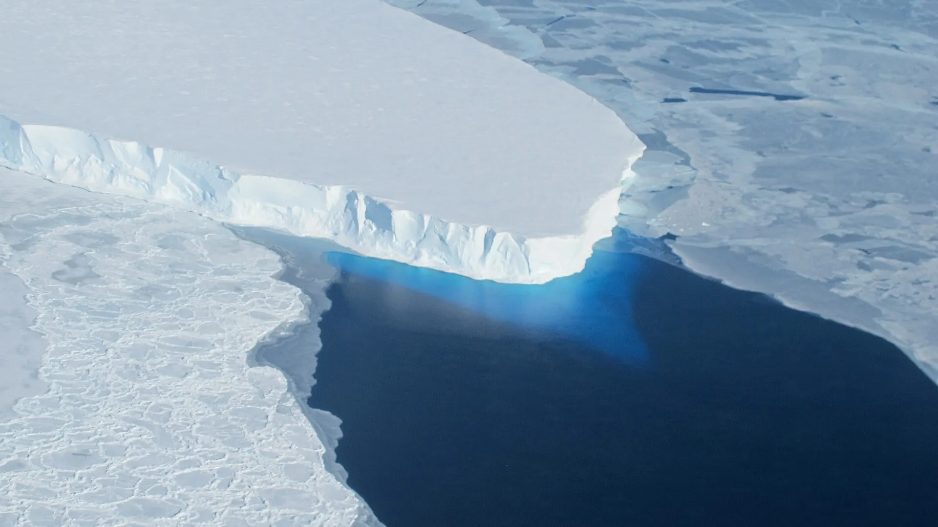The calving front of Thwaites Ice Shelf in West Antarctica photographed from a NASA aircraft, with blue ice visible beneath the water surface
