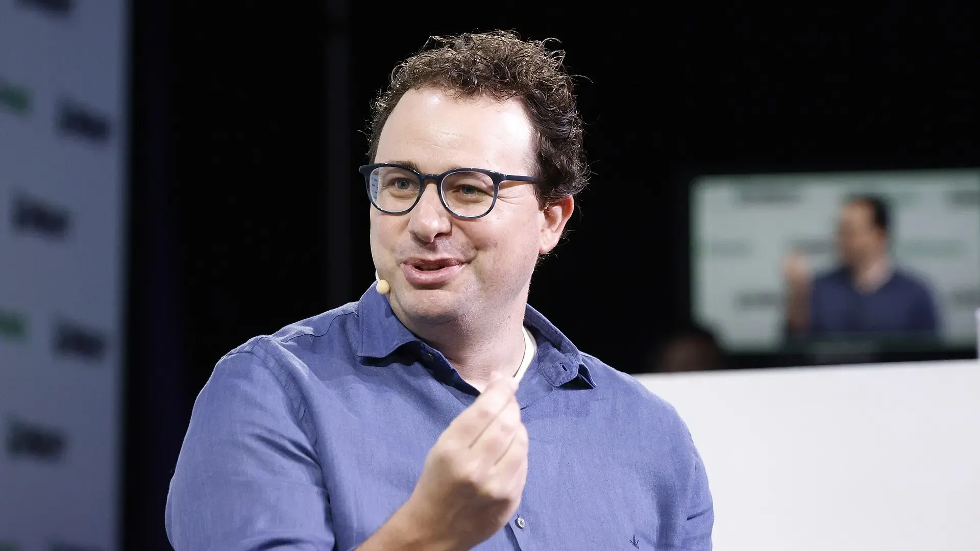 Dario Amodei, wearing glasses and a blue shirt, speaks on stage with a headset microphone in front of a TechCrunch Disrupt backdrop.