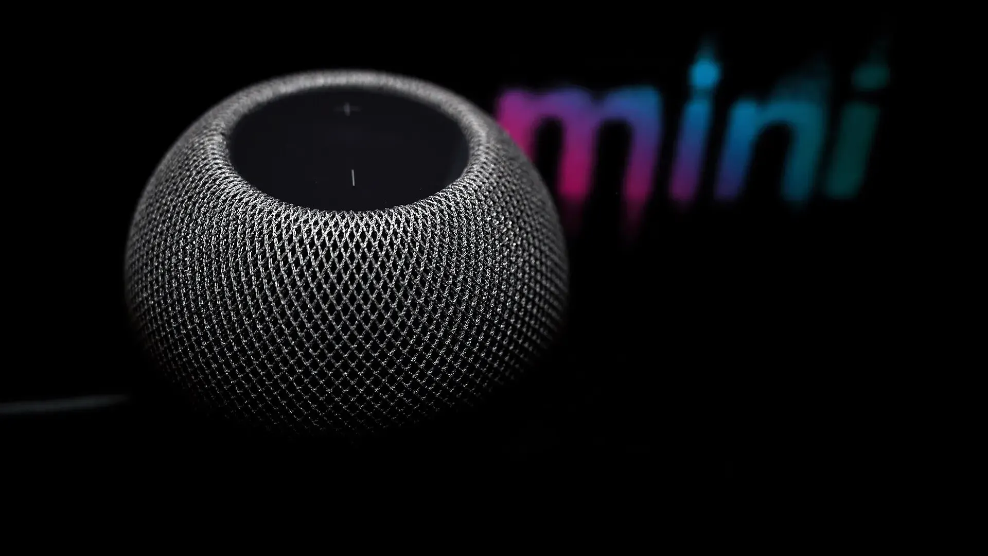A space grey first-generation Apple HomePod mini smart speaker photographed against a black background, with the word mini blurred behind it