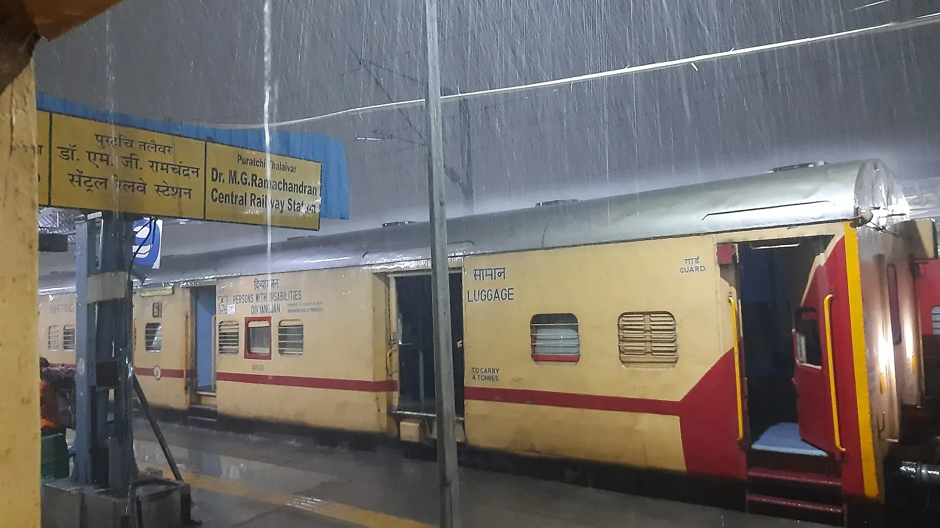 Heavy rain falls on a platform at Chennai Central railway station beside yellow and red train coaches and a bilingual station sign
