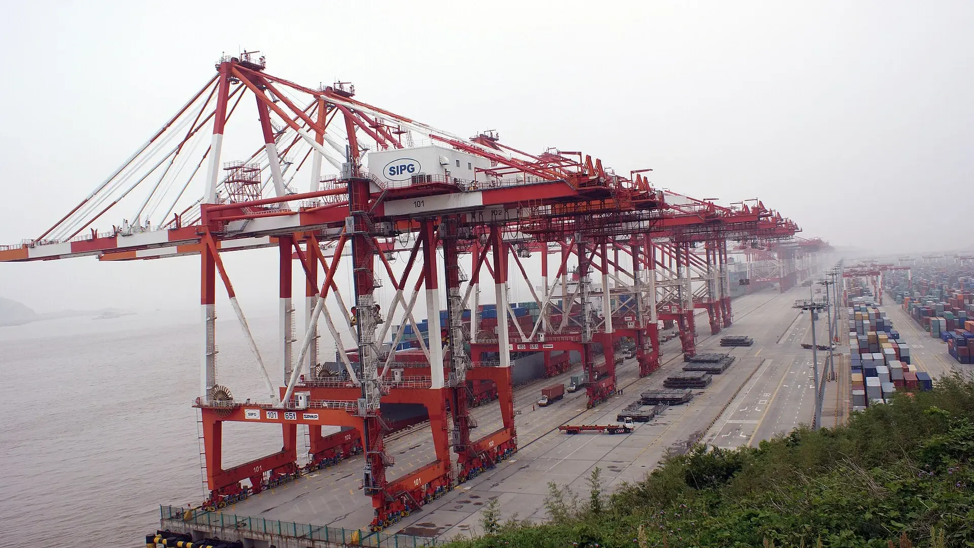 Red and white gantry cranes above stacked shipping containers at the Yangshan deep-water container terminal in Shanghai on a hazy day