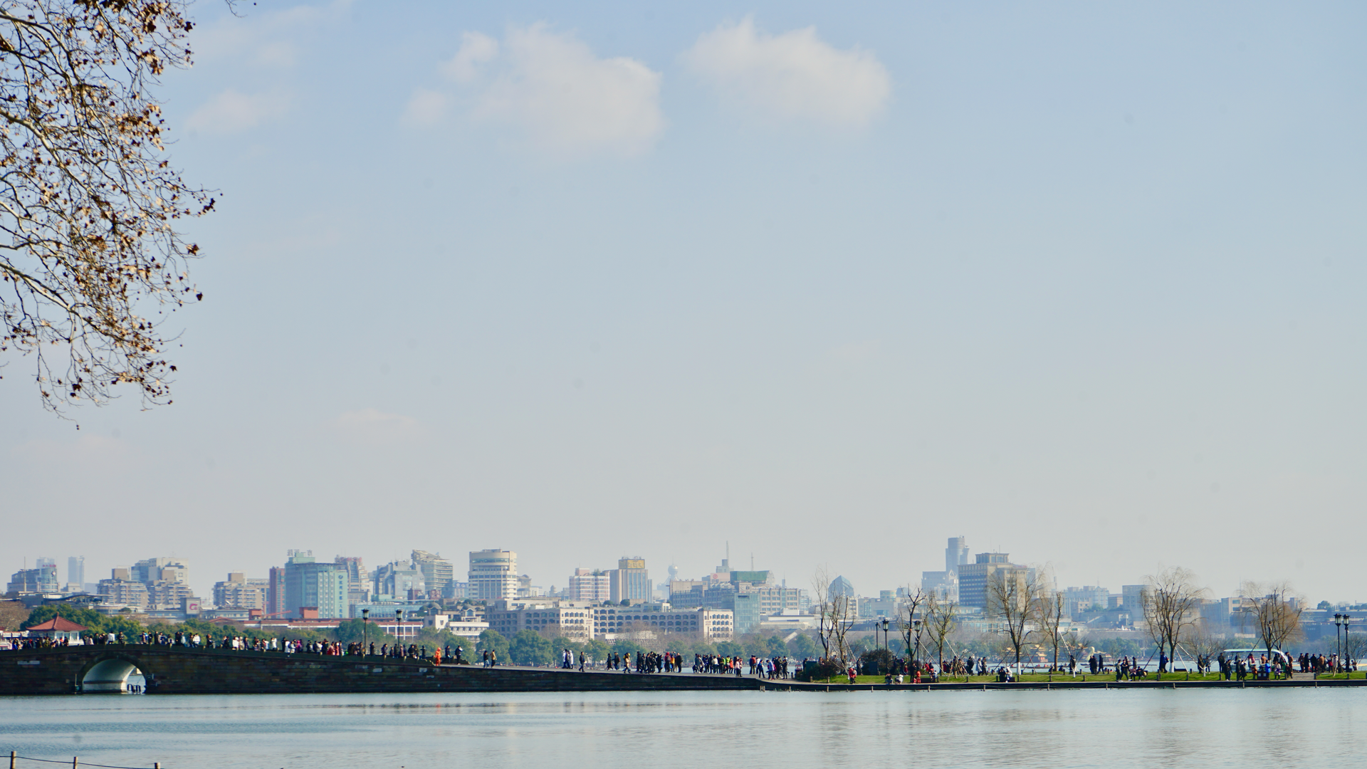 The Hangzhou skyline rising behind the still water of West Lake, with trees along the shoreline in the foreground.