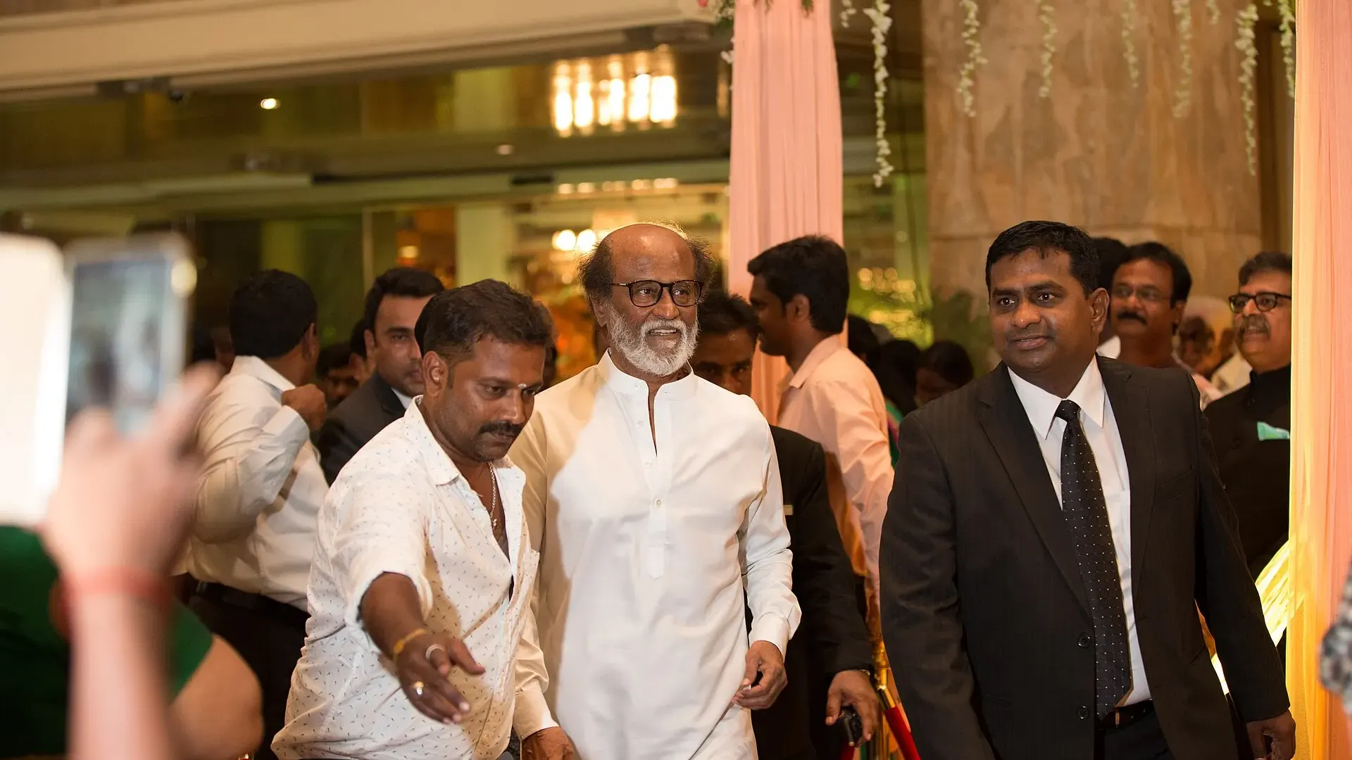 Actor Rajinikanth in a white kurta and dark glasses walking through a crowd at an indoor event in Chennai