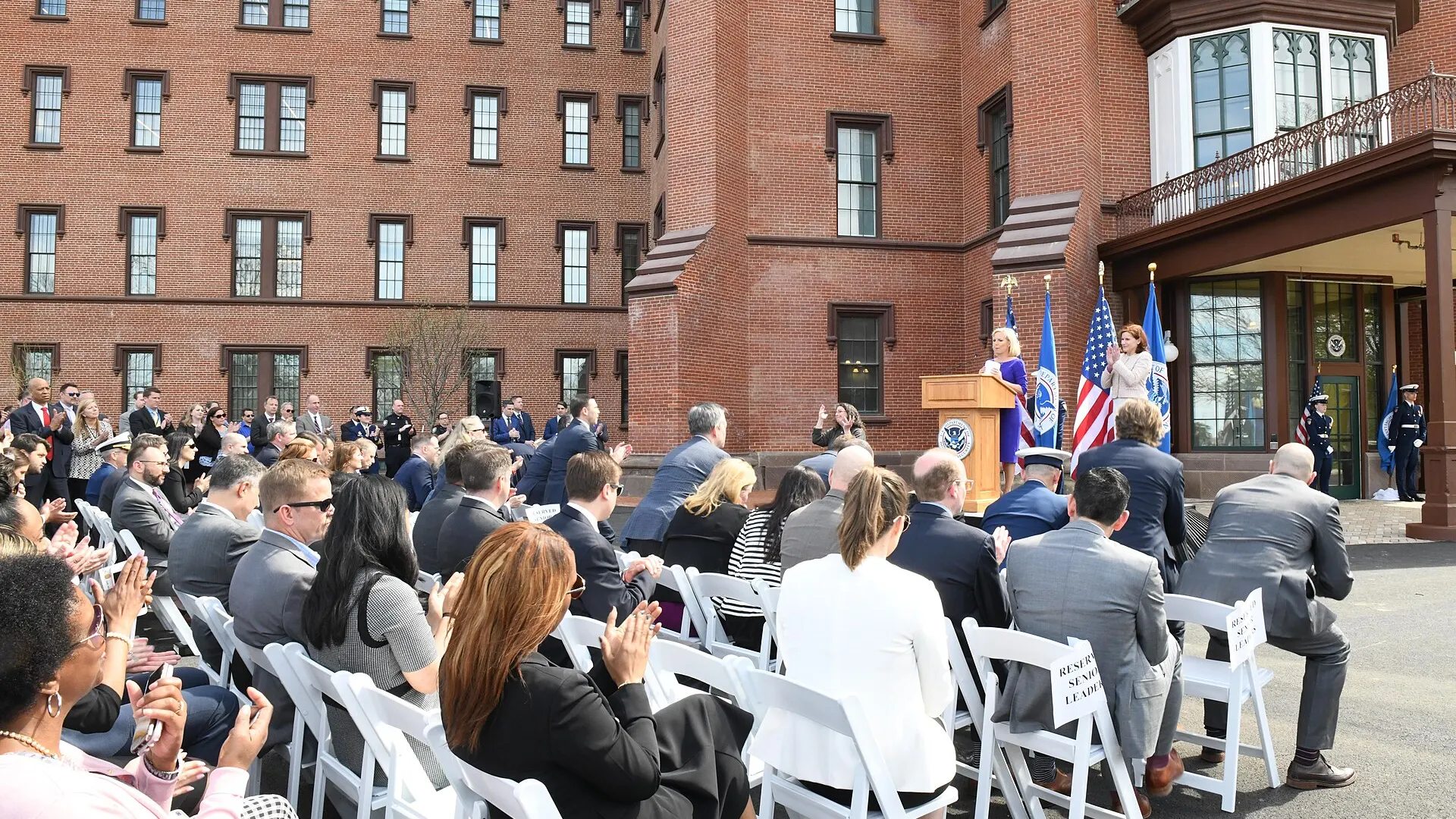 Guests seated in rows of white chairs listen to a speaker at a lectern outside a red-brick building on the St. Elizabeths campus in Washington.