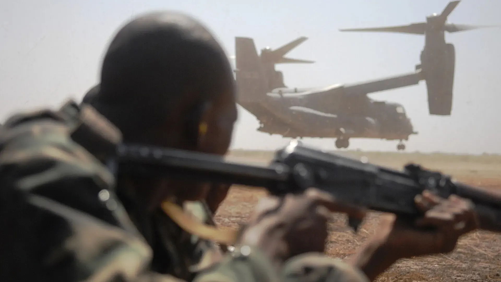 A Malian soldier in camouflage kneels in a defensive position on open ground in Bamako as a military aircraft departs behind him.
