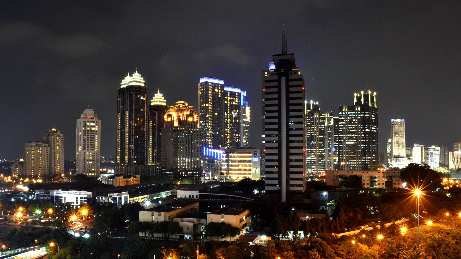 Night view of the lit skyscrapers of the Sudirman Central Business District in Jakarta under a dark sky, with roads and trees in the foreground.