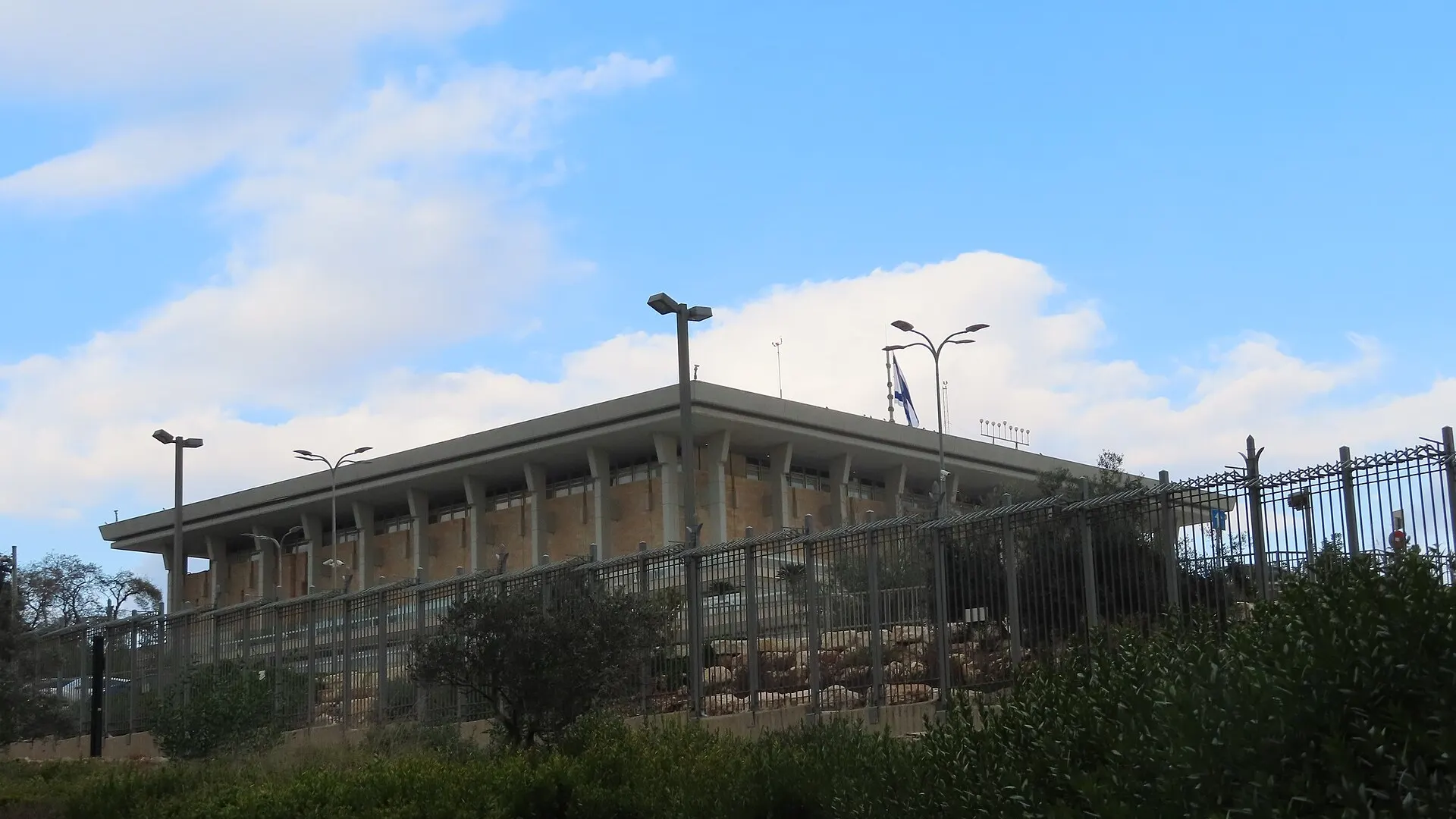 The Knesset building in Jerusalem, a low rectangular stone parliament block set behind lawns and columns under a clear sky