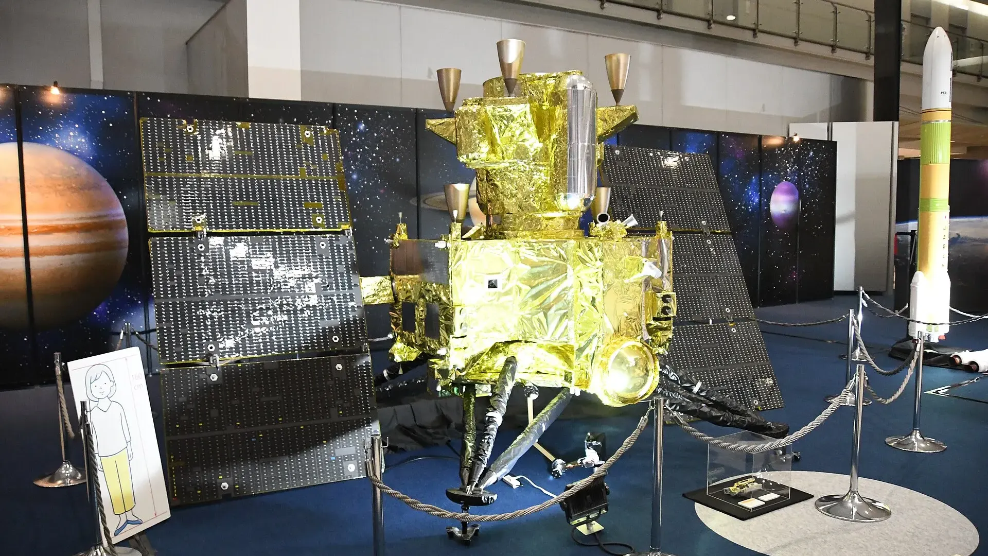 A gold-foil-wrapped scale model of the MMX spacecraft with two dark solar panels and landing legs on display in an exhibition hall, with a model H3 rocket standing to the right.