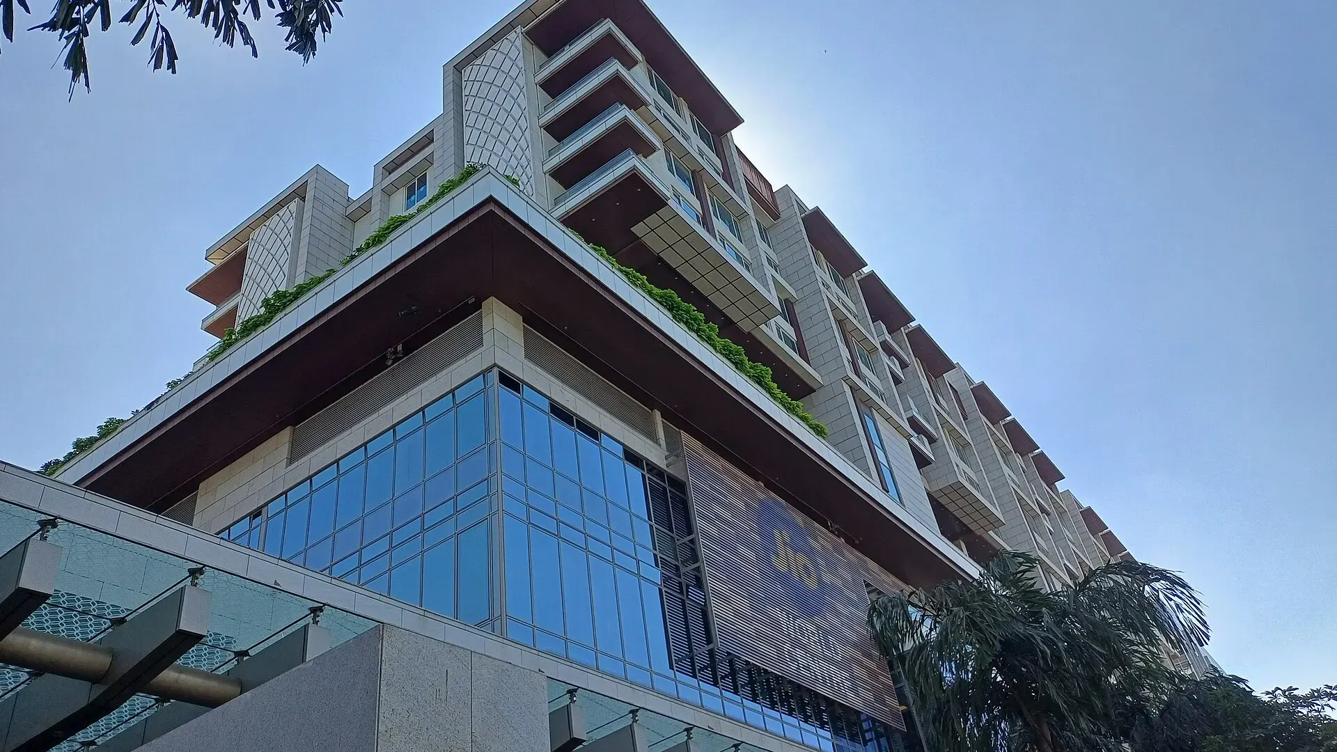 Low-angle view of the glass and stone facade of the Jio World Centre in Mumbai, with Jio World signage, palm trees and blue sky