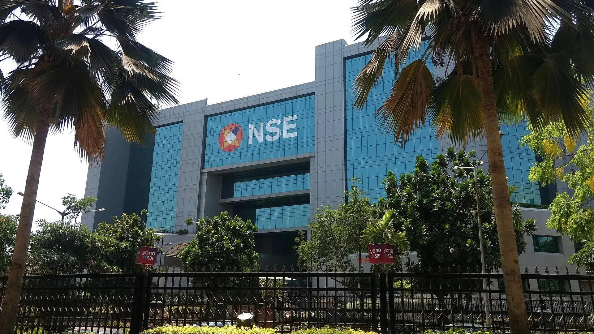 The blue glass facade and NSE sign of the National Stock Exchange building in Mumbai, seen from the street behind palm trees and railings