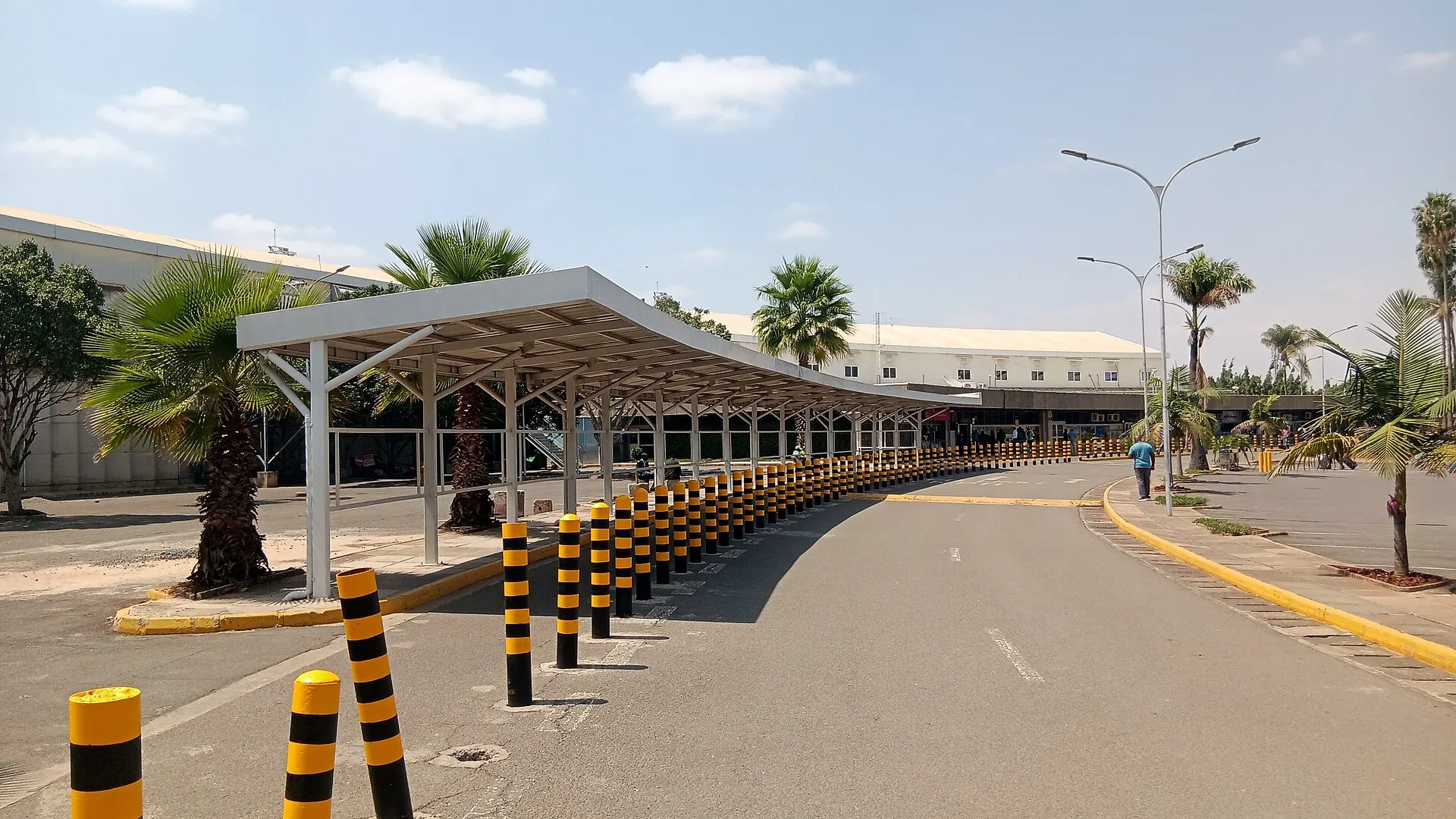 A covered walkway and rows of black and yellow bollards lead to the low white terminal building at Jomo Kenyatta International Airport in Nairobi, with palm trees under a clear sky.