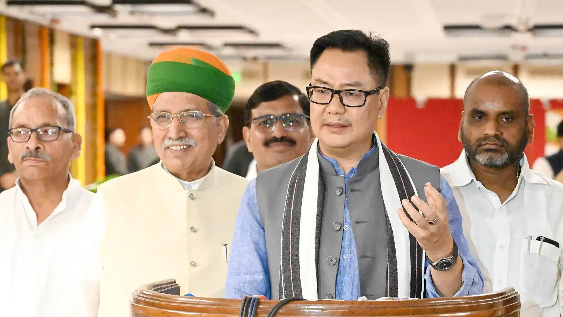 Kiren Rijiju in glasses and a grey waistcoat speaks at a lectern in Parliament House beside Arjun Ram Meghwal in a green and saffron turban