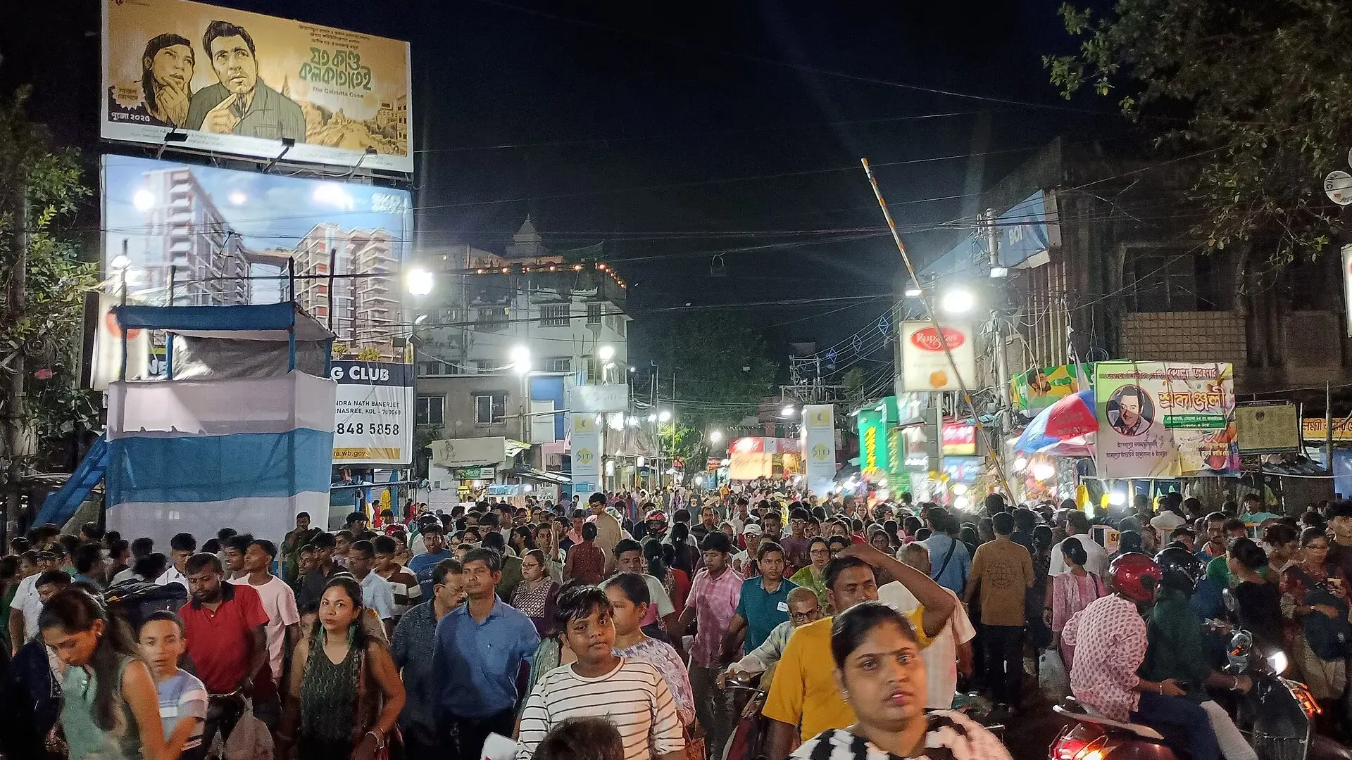 Dense crowd of festival-goers walking along a brightly lit street at night in Behala, Kolkata, during Durga Puja, with billboards overhead.
