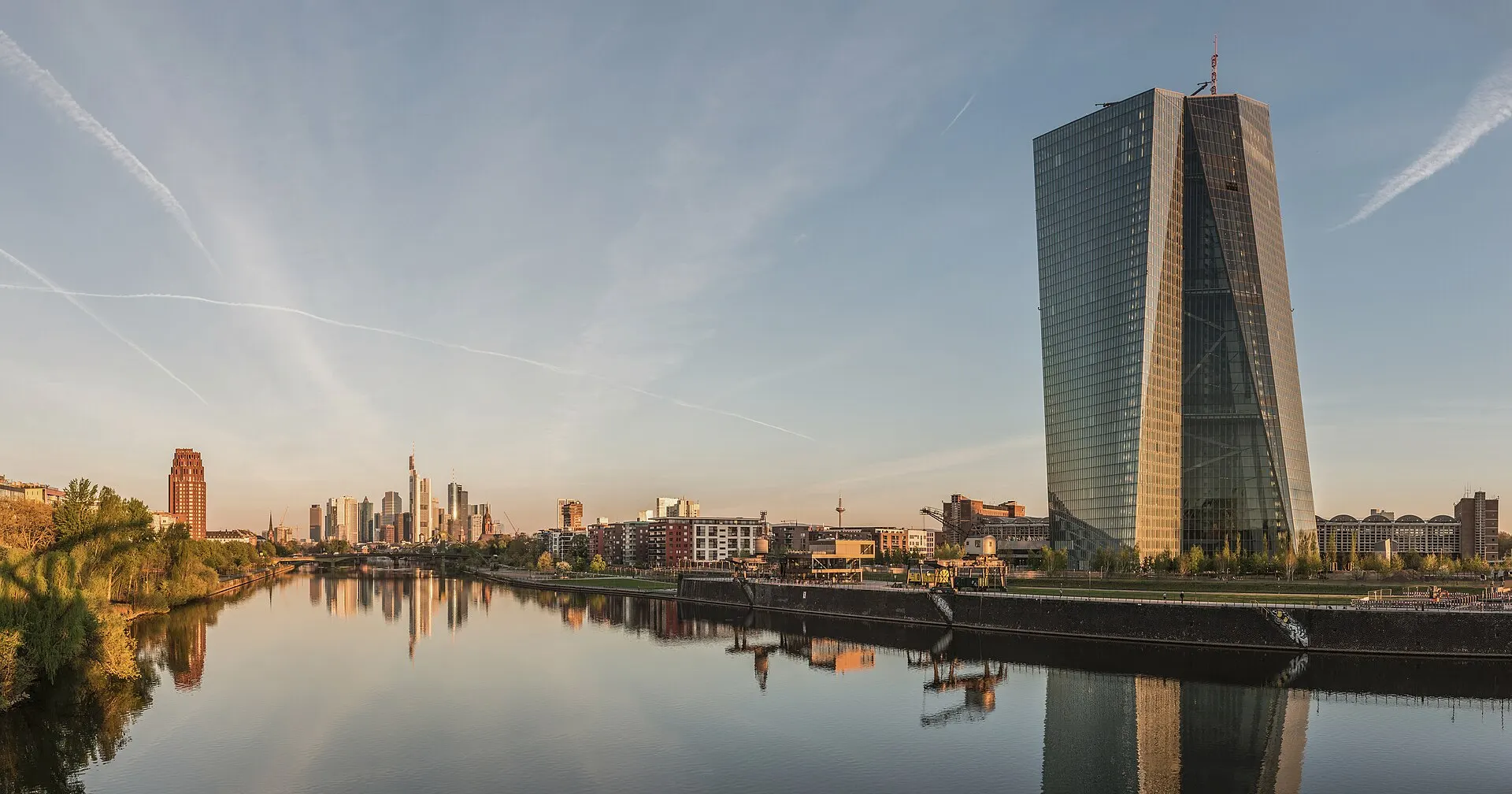 The twin glass towers of the European Central Bank headquarters rise beside the River Main at dawn, with the Frankfurt skyline behind
