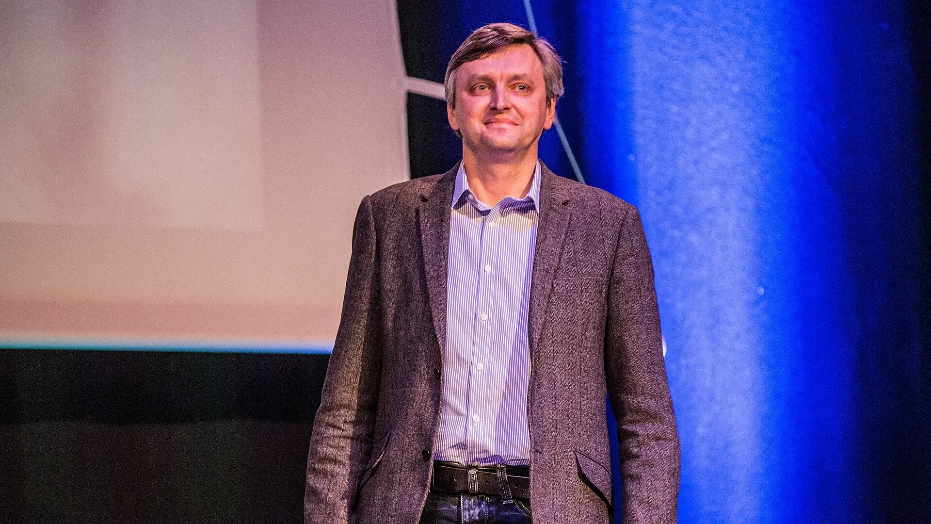 Director Sergei Loznitsa in a grey jacket and striped shirt standing on a cinema stage in front of a blue-lit screen