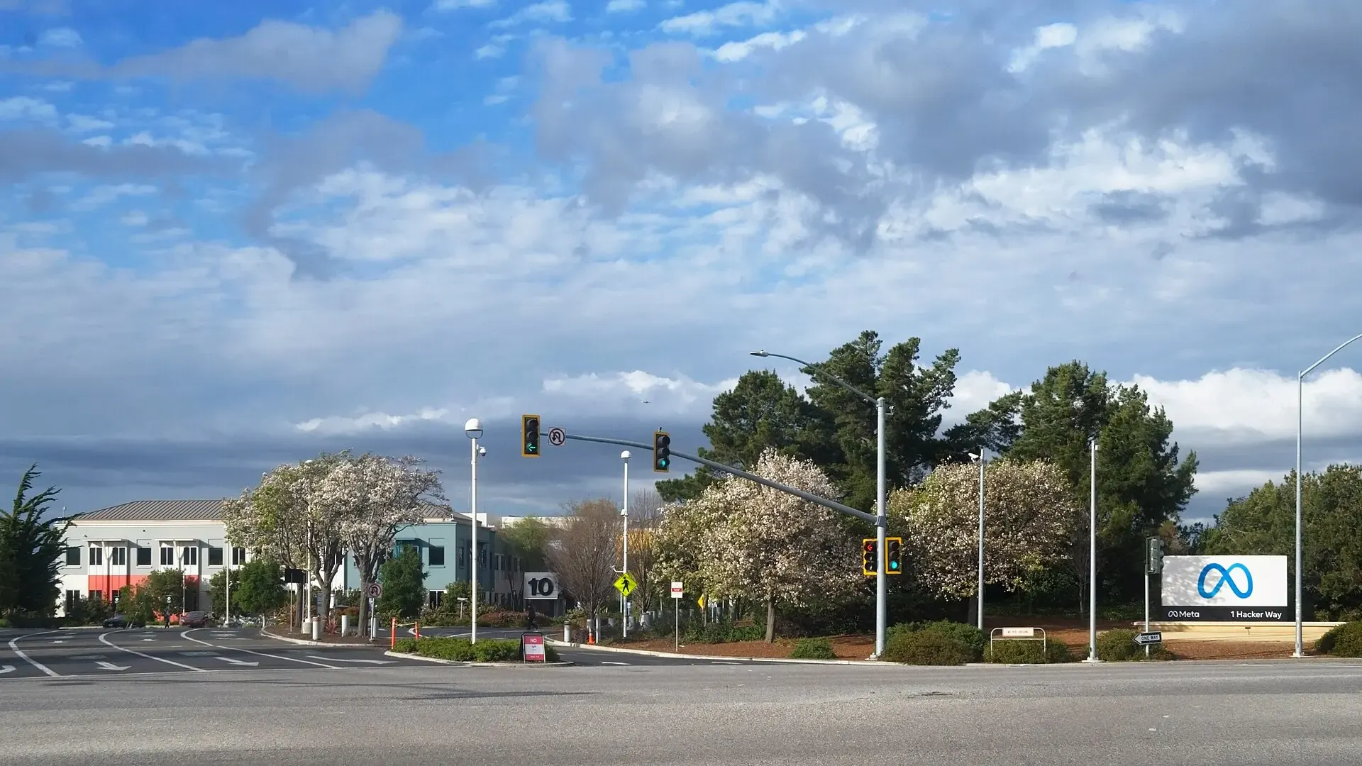The Meta sign at 1 Hacker Way outside the company's Menlo Park, California headquarters, with office buildings and trees behind a road junction.