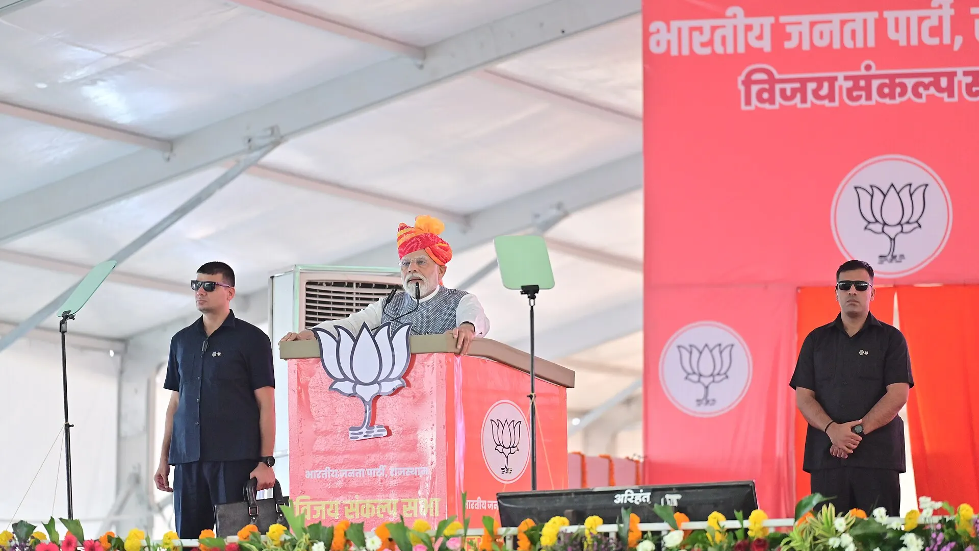 Prime Minister Narendra Modi speaking from a stage to a large crowd at a public rally in India