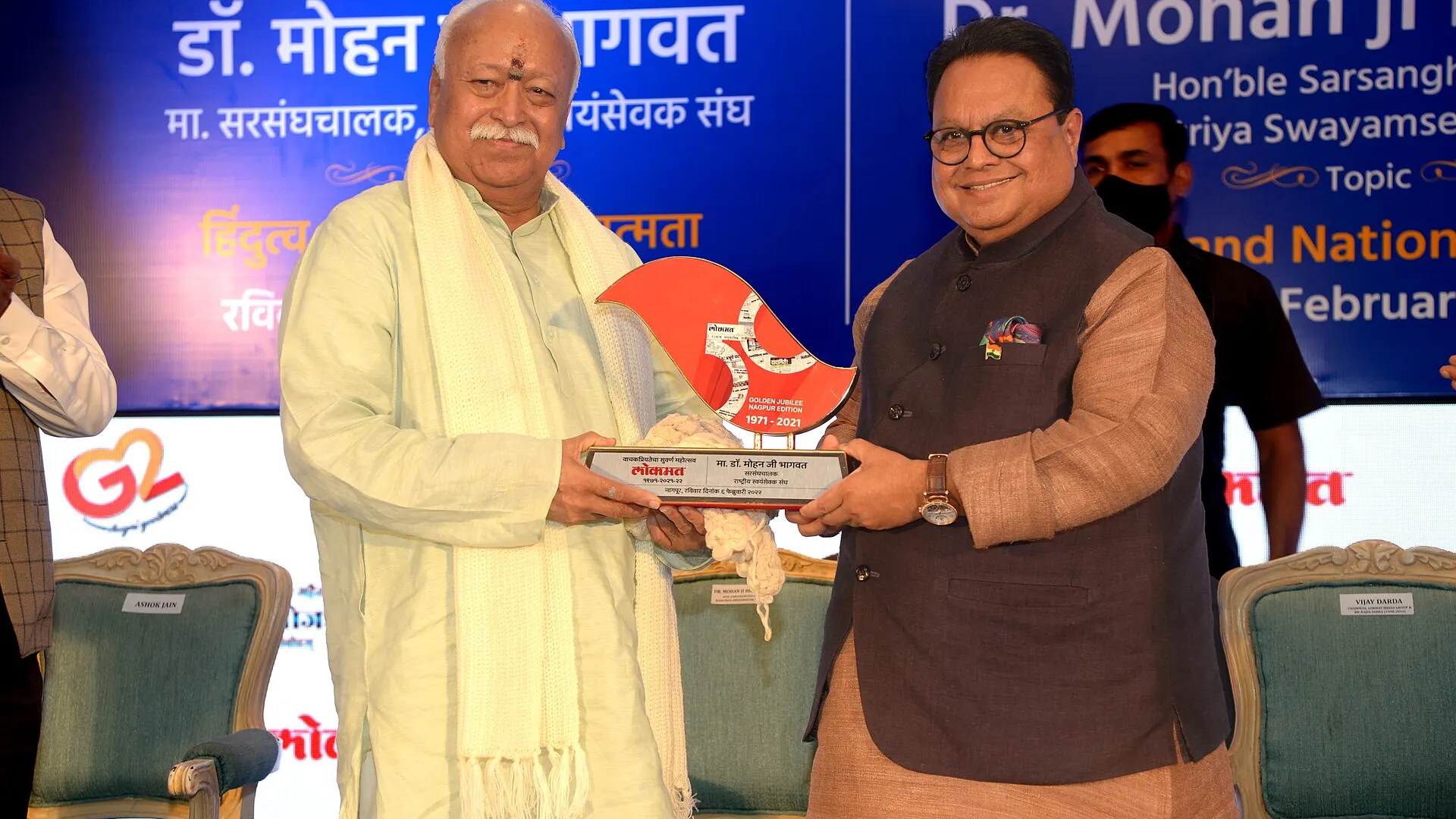 Mohan Bhagwat in a cream kurta and white shawl holds a memento with Vijay Darda, in a dark waistcoat, before a blue stage banner