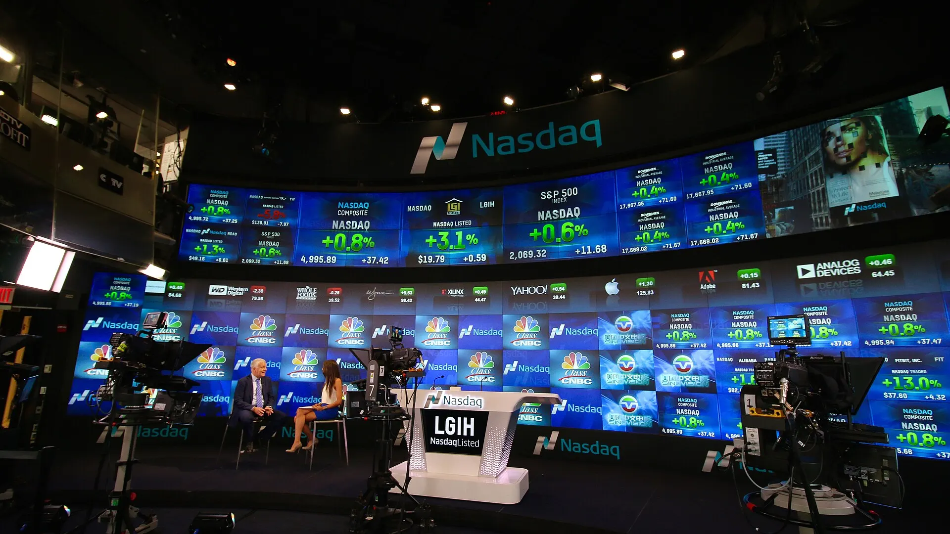 A broadcast studio inside the Nasdaq MarketSite in New York, with a presenter and guest seated in front of curved walls of screens showing stock indexes