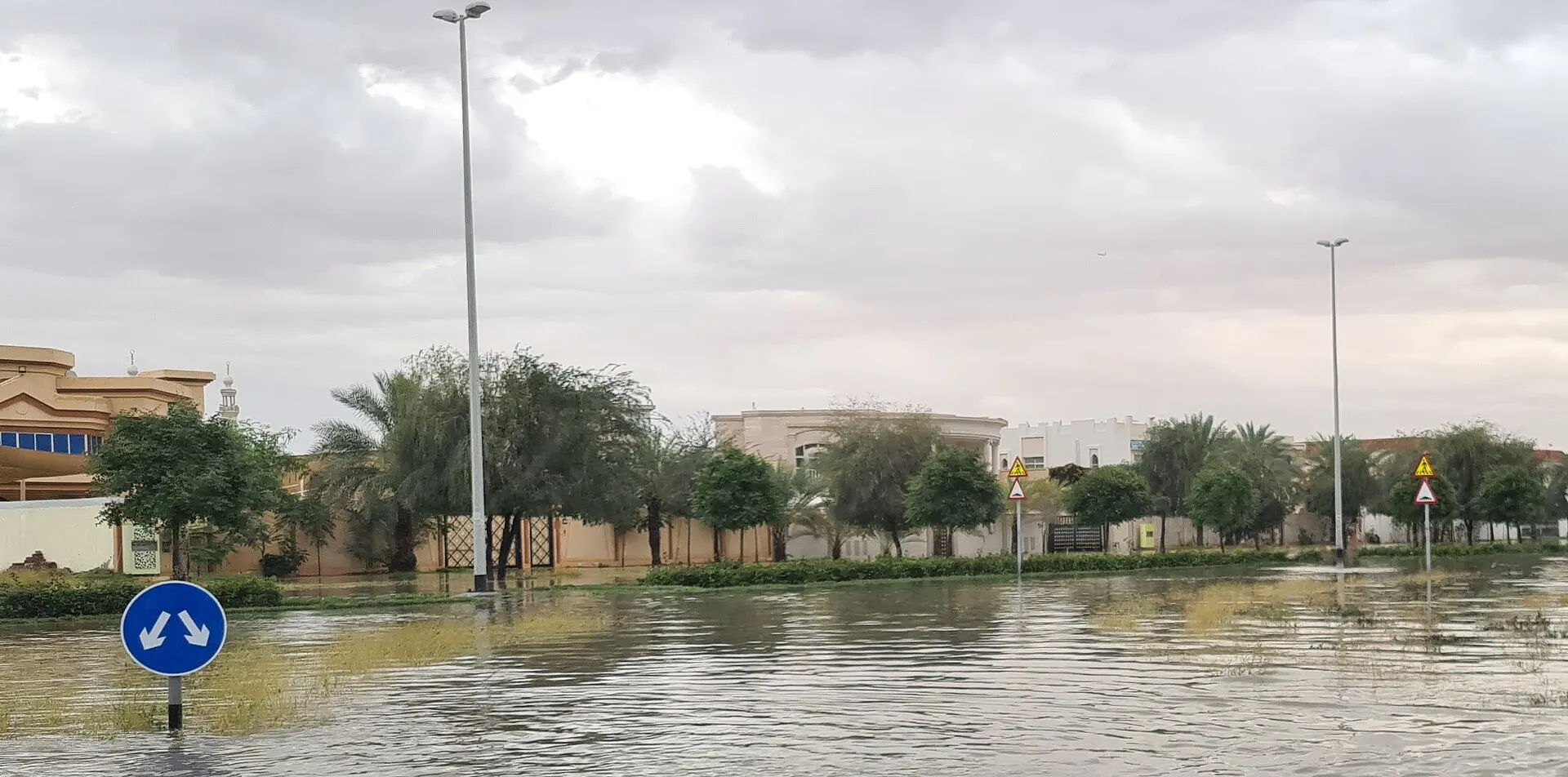 Floodwater covering a residential street in the UAE under grey clouds, with a blue traffic sign, trees and villa walls in April 2024