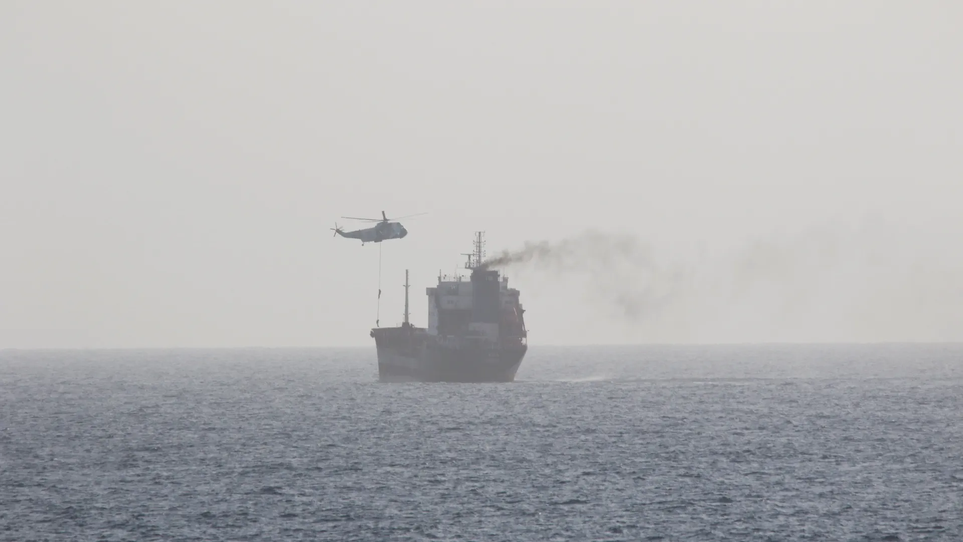 A military helicopter hovers over a tanker at sea in hazy grey light, lowering personnel to the deck by rope as smoke trails from the funnel
