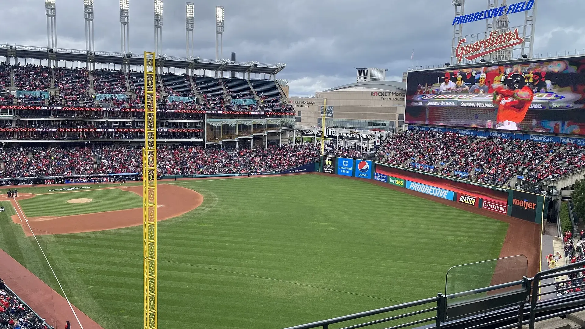 A packed Progressive Field in Cleveland during a postseason baseball game, seen from the upper deck behind home plate with the downtown skyline beyond the outfield.