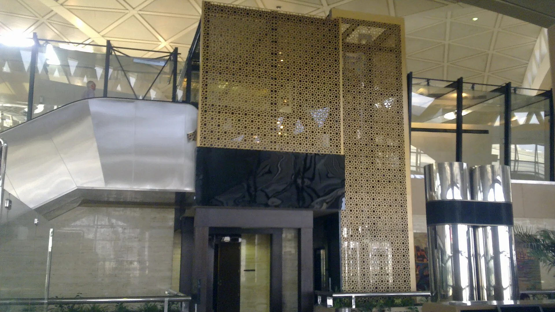 Interior of a passenger terminal at King Khalid International Airport in Riyadh, with a triangular coffered ceiling, escalators and a gilded lattice screen
