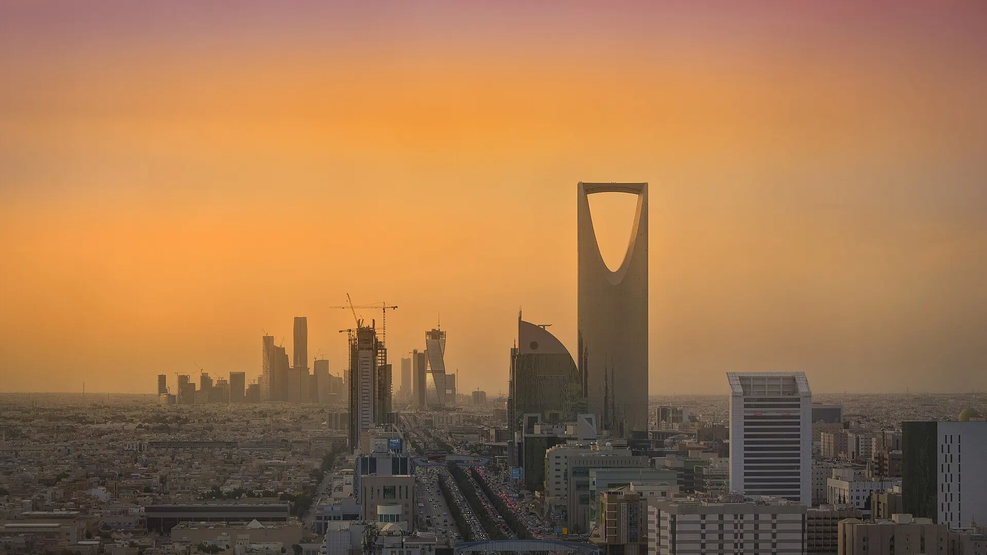 The Kingdom Centre tower rises above central Riyadh at sunset, with the towers and cranes of the King Abdullah Financial District on the horizon