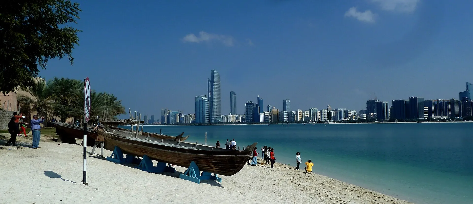 Abu Dhabi's high-rise skyline across turquoise water, with two wooden dhows and visitors on a sandy beach in the foreground
