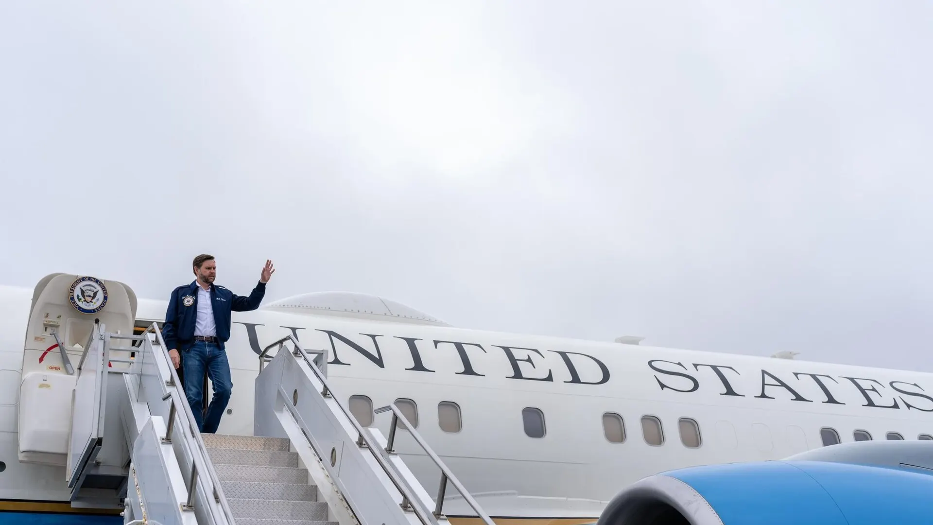 US Vice President JD Vance waves from the top of the airstairs of a United States government aircraft under an overcast sky