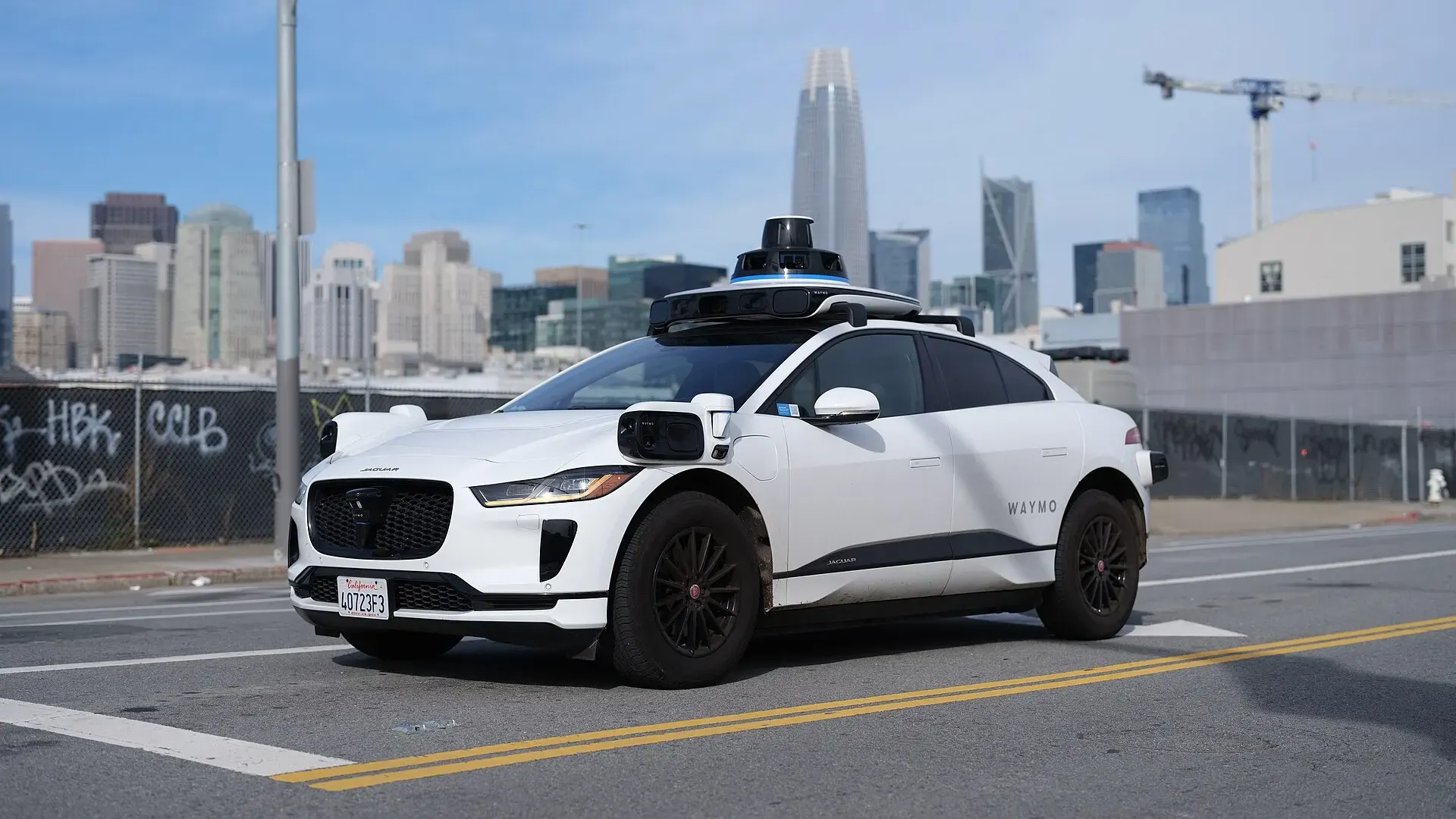A white Waymo Jaguar I-Pace with roof-mounted sensors drives along a San Francisco street with nobody in the driver's seat