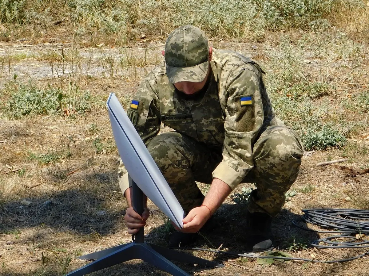 A Ukrainian soldier in camouflage crouches on dry grass while setting up a white Starlink satellite terminal on a tripod.